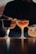 Two cocktails in stem glasses are placed on a reflective surface. One glass contains a dark beverage, while the other holds a lighter-colored drink. A person in dark attire appears in the background, using tools to prepare or garnish the drinks.