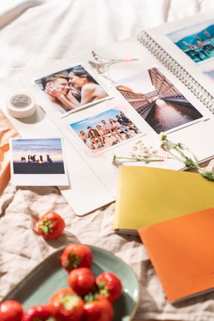 A scrapbook is open with several printed photographs displayed, including a close-up of a couple, a cityscape, a group of people sitting together, and a silhouette of people against a sunset. There are small white flowers and a roll of decorative tape on the scrapbook. Surrounding the scrapbook are strawberries on a plate, two closed notebooks in yellow and orange, and a soft fabric background.