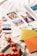 A scrapbook is open with several printed photographs displayed, including a close-up of a couple, a cityscape, a group of people sitting together, and a silhouette of people against a sunset. There are small white flowers and a roll of decorative tape on the scrapbook. Surrounding the scrapbook are strawberries on a plate, two closed notebooks in yellow and orange, and a soft fabric background.