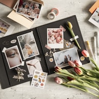 A creatively arranged scrapbook layout featuring various photographs, stickers, and decorative elements on a wooden surface. The open scrapbook displays photos of different subjects including people and flowers, surrounded by vibrant stickers and washi tape. Tulips and small dried flowers add a natural touch to the scene.