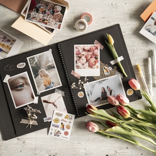 A creatively arranged scrapbook layout featuring various photographs, stickers, and decorative elements on a wooden surface. The open scrapbook displays photos of different subjects including people and flowers, surrounded by vibrant stickers and washi tape. Tulips and small dried flowers add a natural touch to the scene.