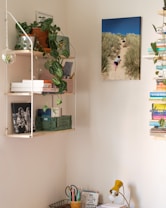 A cozy interior corner with shelves filled with potted plants, books, and assorted objects. A wall has a photo of a sandy path through grass, with people walking in the distance. The workspace has a yellow desk lamp, a basket with colored pens, and various notebooks and office supplies arranged tidily.