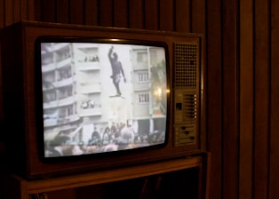 A vintage television set displays a grainy black and white image showing a statue of a man, possibly being toppled in front of large buildings and a crowd. The television is situated in a dimly lit room with wooden paneling.