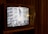 A vintage television set displays a grainy black and white image showing a statue of a man, possibly being toppled in front of large buildings and a crowd. The television is situated in a dimly lit room with wooden paneling.