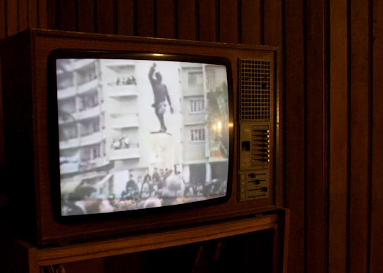 A vintage television set displays a grainy black and white image showing a statue of a man, possibly being toppled in front of large buildings and a crowd. The television is situated in a dimly lit room with wooden paneling.
