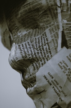 A sculpture or figure is covered in layers of newspaper with visible text in a foreign language. The intricate folds and creases of the paper conform to the contours of a human face, emphasizing the nose and lips, and creating a detailed texture.
