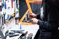 A person is working in a workshop space, holding and adjusting a tool on a black tray. Various tools are organized on a white pegboard to the left, and a bright orange structure, possibly a bicycle frame, is visible in the background. There are several tools and objects laid out on the workbench, including a bike seat and other components.