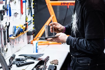 A person is working in a workshop space, holding and adjusting a tool on a black tray. Various tools are organized on a white pegboard to the left, and a bright orange structure, possibly a bicycle frame, is visible in the background. There are several tools and objects laid out on the workbench, including a bike seat and other components.