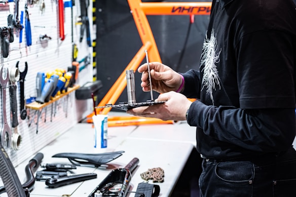 A person is working in a workshop space, holding and adjusting a tool on a black tray. Various tools are organized on a white pegboard to the left, and a bright orange structure, possibly a bicycle frame, is visible in the background. There are several tools and objects laid out on the workbench, including a bike seat and other components.