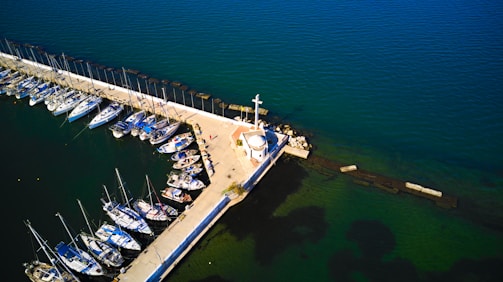 Aerial view of a marina featuring numerous sailboats docked along a pier. A small white lighthouse stands prominently at the pier's end, surrounded by calm, deep blue-green water.
