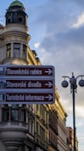 A European street scene featuring an ornate, historic building with a rounded corner tower and decorative architecture. In the foreground, a directional street sign in Czech language points towards several tourist attractions, including the Old Town Hall and a theater. The sky is partly cloudy with a soft, evening light casting shadows.
