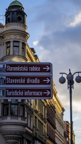 A European street scene featuring an ornate, historic building with a rounded corner tower and decorative architecture. In the foreground, a directional street sign in Czech language points towards several tourist attractions, including the Old Town Hall and a theater. The sky is partly cloudy with a soft, evening light casting shadows.
