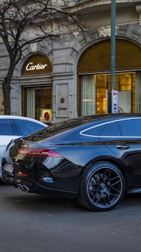 A sleek black luxury car is parked on a street in front of a Cartier store. The vehicle's modern design is accentuated by its glossy finish and stylish wheels. The background features the elegant facade of the store with its arched windows and a prominent Cartier logo. A leafless tree and another white vehicle are also visible.