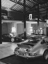 A collection of classic cars positioned in a spacious, industrial-style garage with high ceilings and exposed beams. The cars include a prominently featured vintage Porsche in the foreground with a 'Martini Racing' livery, surrounded by other sports cars on both the ground floor and an elevated platform. Modern chandeliers and neon signs provide contrast to the rustic elements of the space.