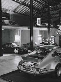 A collection of classic cars positioned in a spacious, industrial-style garage with high ceilings and exposed beams. The cars include a prominently featured vintage Porsche in the foreground with a 'Martini Racing' livery, surrounded by other sports cars on both the ground floor and an elevated platform. Modern chandeliers and neon signs provide contrast to the rustic elements of the space.