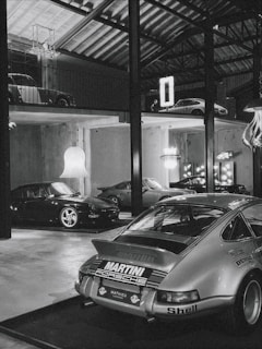 A collection of classic cars positioned in a spacious, industrial-style garage with high ceilings and exposed beams. The cars include a prominently featured vintage Porsche in the foreground with a 'Martini Racing' livery, surrounded by other sports cars on both the ground floor and an elevated platform. Modern chandeliers and neon signs provide contrast to the rustic elements of the space.