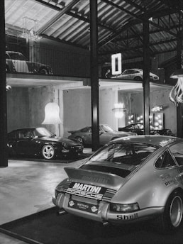A collection of classic cars positioned in a spacious, industrial-style garage with high ceilings and exposed beams. The cars include a prominently featured vintage Porsche in the foreground with a 'Martini Racing' livery, surrounded by other sports cars on both the ground floor and an elevated platform. Modern chandeliers and neon signs provide contrast to the rustic elements of the space.