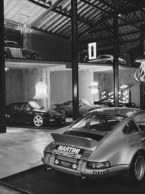 A collection of classic cars positioned in a spacious, industrial-style garage with high ceilings and exposed beams. The cars include a prominently featured vintage Porsche in the foreground with a 'Martini Racing' livery, surrounded by other sports cars on both the ground floor and an elevated platform. Modern chandeliers and neon signs provide contrast to the rustic elements of the space.