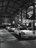 Several classic race cars are on display inside a large, well-lit industrial-style garage or showroom. The cars have distinctive racing numbers and sponsorship decals. The room has high ceilings with exposed metal beams and hanging light fixtures. A decorative chandelier hangs from the ceiling, adding a touch of elegance to the industrial space.