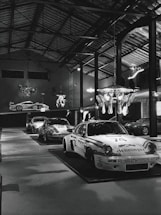 Several classic race cars are on display inside a large, well-lit industrial-style garage or showroom. The cars have distinctive racing numbers and sponsorship decals. The room has high ceilings with exposed metal beams and hanging light fixtures. A decorative chandelier hangs from the ceiling, adding a touch of elegance to the industrial space.