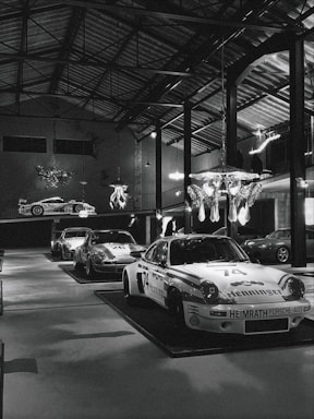 Several classic race cars are on display inside a large, well-lit industrial-style garage or showroom. The cars have distinctive racing numbers and sponsorship decals. The room has high ceilings with exposed metal beams and hanging light fixtures. A decorative chandelier hangs from the ceiling, adding a touch of elegance to the industrial space.