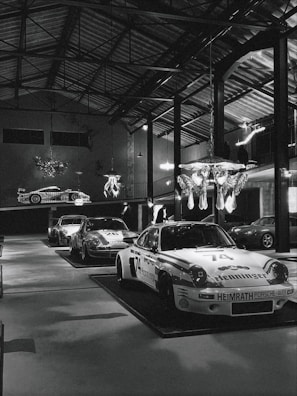 Several classic race cars are on display inside a large, well-lit industrial-style garage or showroom. The cars have distinctive racing numbers and sponsorship decals. The room has high ceilings with exposed metal beams and hanging light fixtures. A decorative chandelier hangs from the ceiling, adding a touch of elegance to the industrial space.