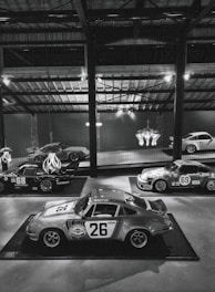 A collection of vintage racing cars displayed in a dimly lit, industrial-style showroom. Prominent is a classic car with the number 26 and 'Martini Racing' branding, surrounded by other vintage cars, some with numbers 68 and 69, showcasing retro racing decals. The ambient lighting is mellow, with overhead beams and a chandelier adding to the aesthetic.
