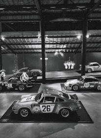 A collection of vintage racing cars displayed in a dimly lit, industrial-style showroom. Prominent is a classic car with the number 26 and 'Martini Racing' branding, surrounded by other vintage cars, some with numbers 68 and 69, showcasing retro racing decals. The ambient lighting is mellow, with overhead beams and a chandelier adding to the aesthetic.