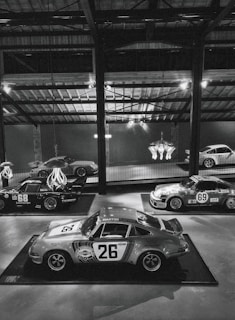A collection of vintage racing cars displayed in a dimly lit, industrial-style showroom. Prominent is a classic car with the number 26 and 'Martini Racing' branding, surrounded by other vintage cars, some with numbers 68 and 69, showcasing retro racing decals. The ambient lighting is mellow, with overhead beams and a chandelier adding to the aesthetic.