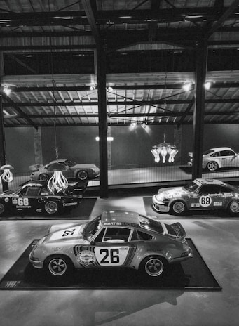 A collection of vintage racing cars displayed in a dimly lit, industrial-style showroom. Prominent is a classic car with the number 26 and 'Martini Racing' branding, surrounded by other vintage cars, some with numbers 68 and 69, showcasing retro racing decals. The ambient lighting is mellow, with overhead beams and a chandelier adding to the aesthetic.