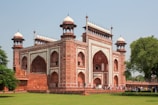 A large and ornate red sandstone and marble building with intricate architectural details, including arched entrances and decorative towers. It features geometric and floral patterns along its surfaces. A group of people is gathered near the entrance, and the building is surrounded by lush green lawns and trees under a clear blue sky.