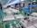 The interior of a factory or assembly line is visible, with several workstations lined up neatly. Each station contains white plastic enclosures with electronic components inside, suggesting an electronic manufacturing or assembly process. Overhead lighting and blue tool boards are present, with some orange coiled cords hanging down.