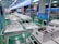 The interior of a factory or assembly line is visible, with several workstations lined up neatly. Each station contains white plastic enclosures with electronic components inside, suggesting an electronic manufacturing or assembly process. Overhead lighting and blue tool boards are present, with some orange coiled cords hanging down.