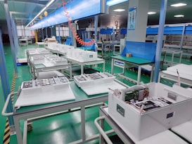 The interior of a factory or assembly line is visible, with several workstations lined up neatly. Each station contains white plastic enclosures with electronic components inside, suggesting an electronic manufacturing or assembly process. Overhead lighting and blue tool boards are present, with some orange coiled cords hanging down.