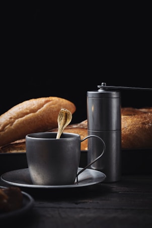 A minimalist composition featuring a metal cup and saucer with a gold spoon inside. Beside the cup is a sleek metallic salt or pepper grinder. In the background, several loaves of crusty bread are partially visible, all set against a dark backdrop, creating a sophisticated and moody atmosphere.