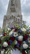 A tall stone monument with intricate carvings of human figures stands in the background, partially covered by a large, vibrant floral arrangement in the foreground. The flowers include red roses, white daisies, and blue blossoms, set against lush green leaves. The sky is overcast, adding a somber and reflective atmosphere to the scene.