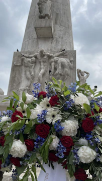 A tall stone monument with intricate carvings of human figures stands in the background, partially covered by a large, vibrant floral arrangement in the foreground. The flowers include red roses, white daisies, and blue blossoms, set against lush green leaves. The sky is overcast, adding a somber and reflective atmosphere to the scene.