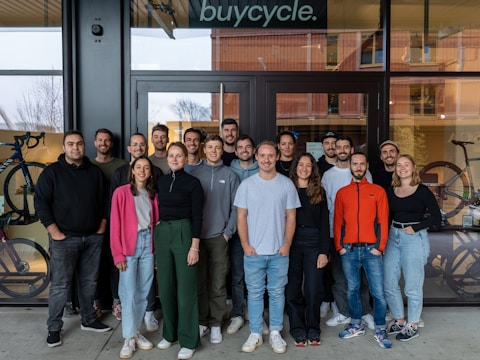 A group of people standing together in front of a glass door with the word 'buycycle' above them, possibly indicating the entrance to a store or office. They are dressed in casual attire, with a visible bicycle in the background on both sides of the door.