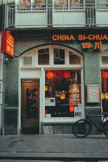 A cozy Chinese restaurant with a green brick facade and orange signage that reads 'China Si-Chuan.' Red lanterns are visible through the windows, adding a warm glow. A menu displayed outside features various dishes. A black bicycle is parked beside the entrance.