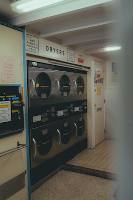 A laundromat interior with multiple industrial-grade dryers stacked in two rows against the wall. There's a vending machine for tokens on the left side and several signs with instructions, including one warning against putting rubber or plastic items in the dryers. The flooring is a mix of tiled and linoleum sections, and the walls are adorned with various informational notices.