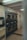 A laundromat interior with multiple industrial-grade dryers stacked in two rows against the wall. There's a vending machine for tokens on the left side and several signs with instructions, including one warning against putting rubber or plastic items in the dryers. The flooring is a mix of tiled and linoleum sections, and the walls are adorned with various informational notices.