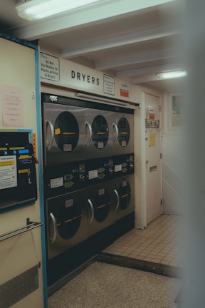 A laundromat interior with multiple industrial-grade dryers stacked in two rows against the wall. There's a vending machine for tokens on the left side and several signs with instructions, including one warning against putting rubber or plastic items in the dryers. The flooring is a mix of tiled and linoleum sections, and the walls are adorned with various informational notices.