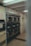 A laundromat interior with multiple industrial-grade dryers stacked in two rows against the wall. There's a vending machine for tokens on the left side and several signs with instructions, including one warning against putting rubber or plastic items in the dryers. The flooring is a mix of tiled and linoleum sections, and the walls are adorned with various informational notices.