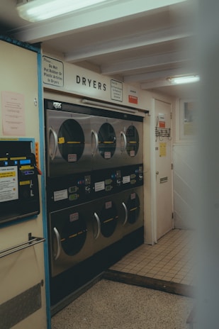 A laundromat interior with multiple industrial-grade dryers stacked in two rows against the wall. There's a vending machine for tokens on the left side and several signs with instructions, including one warning against putting rubber or plastic items in the dryers. The flooring is a mix of tiled and linoleum sections, and the walls are adorned with various informational notices.
