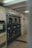 A laundromat interior with multiple industrial-grade dryers stacked in two rows against the wall. There's a vending machine for tokens on the left side and several signs with instructions, including one warning against putting rubber or plastic items in the dryers. The flooring is a mix of tiled and linoleum sections, and the walls are adorned with various informational notices.