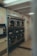 A laundromat interior with multiple industrial-grade dryers stacked in two rows against the wall. There's a vending machine for tokens on the left side and several signs with instructions, including one warning against putting rubber or plastic items in the dryers. The flooring is a mix of tiled and linoleum sections, and the walls are adorned with various informational notices.