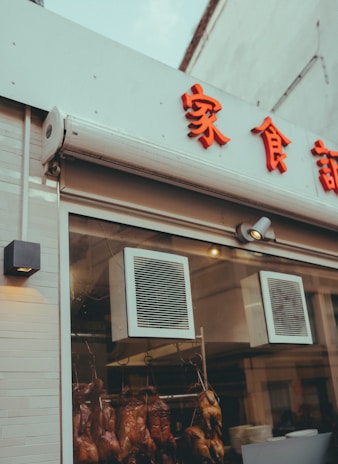 A restaurant window display with roasted ducks hanging inside. The exterior features red Chinese characters on a white background, underlined by two ventilators situated above the window. The setting suggests an Asian eatery specializing in duck dishes.