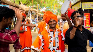 A group of people in vibrant traditional attire, including bright orange turbans and robes, are participating in a lively celebration. One individual is holding a tambourine, while others around him appear to be dancing and enjoying festive music. The setting is an outdoor cultural fair with colorful banners and stalls in the background.