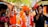 A group of people in vibrant traditional attire, including bright orange turbans and robes, are participating in a lively celebration. One individual is holding a tambourine, while others around him appear to be dancing and enjoying festive music. The setting is an outdoor cultural fair with colorful banners and stalls in the background.