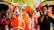 A group of people in vibrant traditional attire, including bright orange turbans and robes, are participating in a lively celebration. One individual is holding a tambourine, while others around him appear to be dancing and enjoying festive music. The setting is an outdoor cultural fair with colorful banners and stalls in the background.